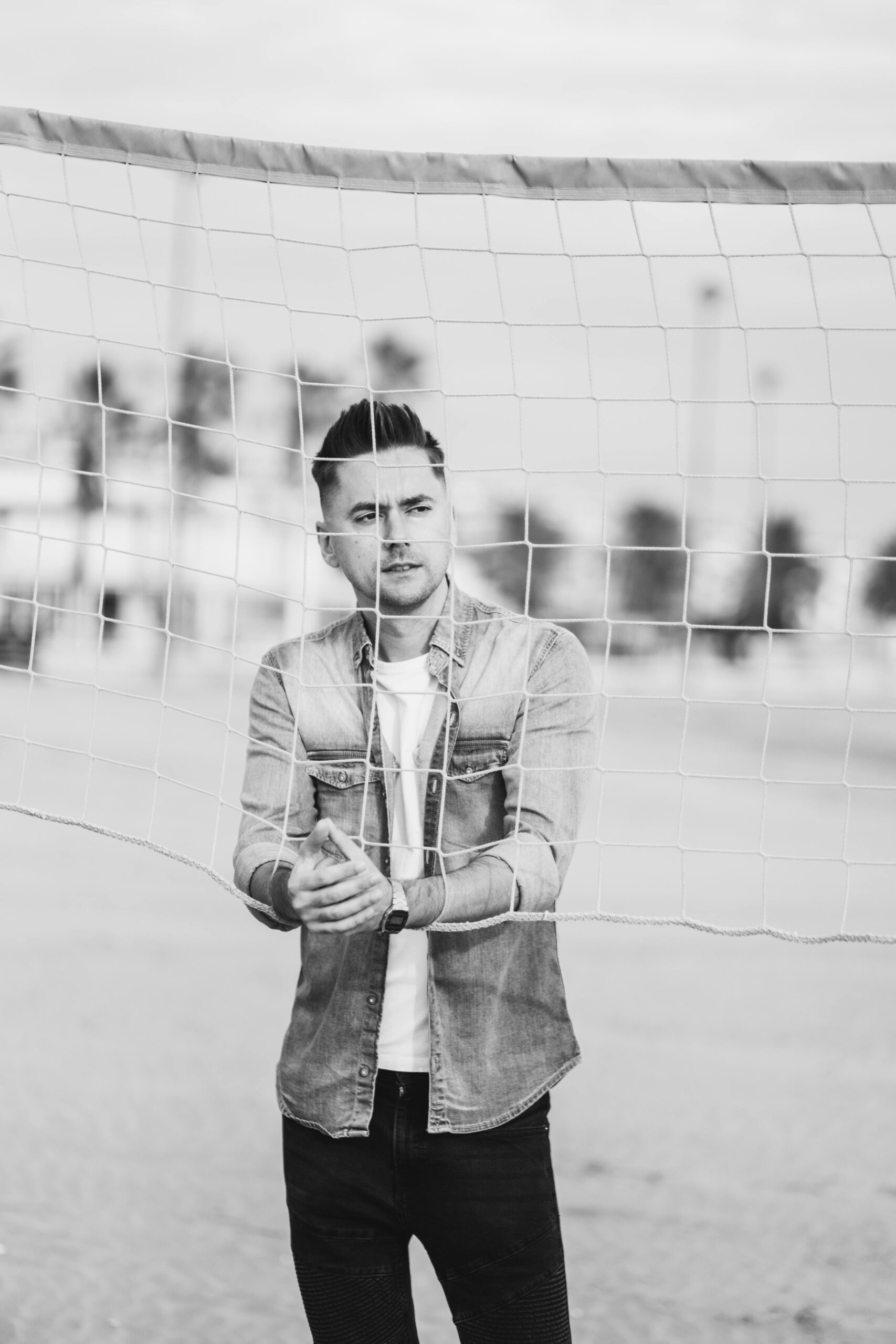 Stylish young man stands behind a volleyball net on a beach, captured in black and white.